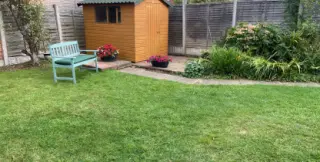 Abri de jardin en bois couvert de plaques bitumées vertes, au fond d'un jardin engazonné