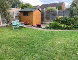 Abri de jardin en bois couvert de plaques bitumées vertes, au fond d'un jardin engazonné