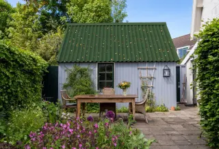 Abri de jardin au toit en plaques bitumées vertes, entouré de haies et de plantes grimpantes