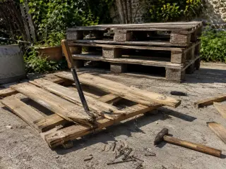 Pile de palettes en bois de récupération empilées dans un jardin avec une palette en cours de démontage au premier plan, pied-de-biche et marteau de charpentier posés sur la dalle