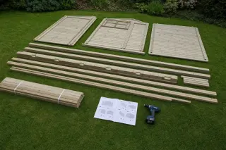 Pièces d'ossature en bois pin pré-percées posées sur pelouse, visseuse électrique noire à côté, instructions de montage A4 ouvertes au sol, bottes en caoutchouc verte plus loin