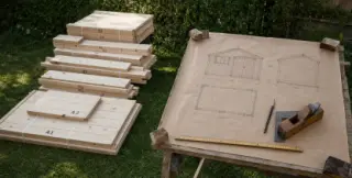 À gauche les pièces préfabriquées d'un abri en kit empilées sur pelouse, à droite un plan d'abri dessiné à la main sur papier kraft posé sur tréteaux avec outils traditionnels
