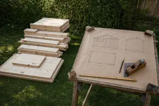 À gauche les pièces préfabriquées d'un abri en kit empilées sur pelouse, à droite un plan d'abri dessiné à la main sur papier kraft posé sur tréteaux avec outils traditionnels