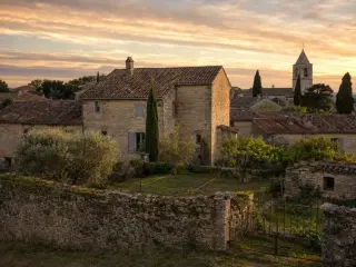 Vue d'ensemble d'un village français historique du sud avec maisons en pierre apparente couleur miel et toitures en tuile rouge traditionnelle, clocher d'église en arrière-plan, ciel orangé de fin d'après-midi