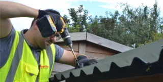 Bricoleur vissant en sommet d'onde une plaque bitumée ondulée ONDULINE EASYFIX® sur la charpente bois d'un abri de jardin en cours de construction