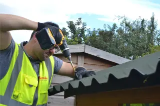 Bricoleur vissant en sommet d'onde une plaque bitumée ondulée ONDULINE EASYFIX® sur la charpente bois d'un abri de jardin en cours de construction