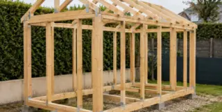 Ossature bois d'un abri de jardin en cours de montage : poteaux, sablières et chevrons assemblés par équerres galvanisées sur des plots béton dans un jardin français