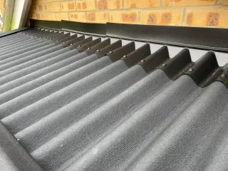 Raccordement de la toiture d'un abri adossé contre un mur de briques : le solin épouse toute l'onde des plaques bitumées ONDULINE anthracite et pas seulement leur sommet, avant de remonter contre le mur, fixations en sommet d'onde