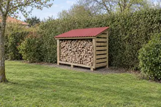 Bûcher en bois à parois à claire-voie rempli de bûches, toit monopente en plaques ondulées rouges, vu de loin devant une haie