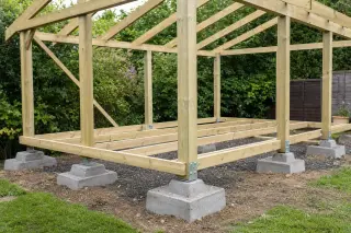 Ossature bois d'un petit abri utilitaire en construction, surélevée sur plots béton, montants verticaux et chevrons assemblés, platines d'ancrage au sol visibles avant pose de la couverture