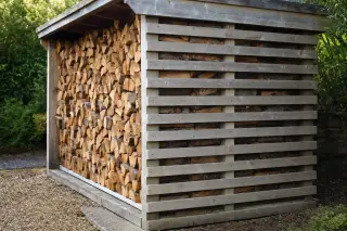 Paroi latérale d'un abri à bois en claire-voie : planches horizontales espacées régulièrement laissant passer l'air, bûches visibles derrière, façade avant ouverte