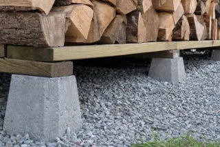 Base d'un abri à bois surélevée sur plots béton et lambourdes en bois traité, avec espace d'air sous la première rangée de bûches, sur un lit de gravier