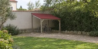Abri à vélos ouvert en bois à toit monopente en plaques ondulées rouges, deux vélos dessous, vu de loin contre un mur de jardin
