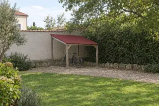 Abri à vélos ouvert en bois à toit monopente en plaques ondulées rouges, deux vélos dessous, vu de loin contre un mur de jardin