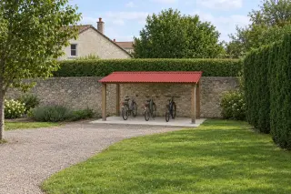 Abri à vélos ouvert en bois, toit monopente en plaques ondulées rouges, quatre vélos rangés côte à côte sur une dalle, vu de loin devant un mur de pierre